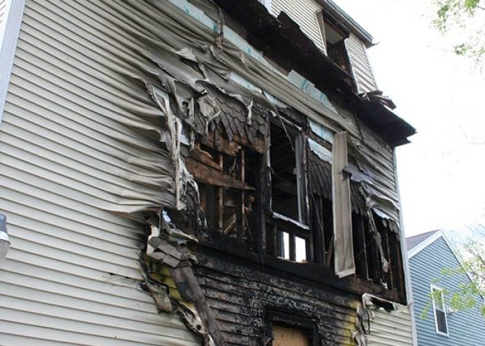 Fire-damaged exterior wall with burned siding and exposed framing around a window opening.